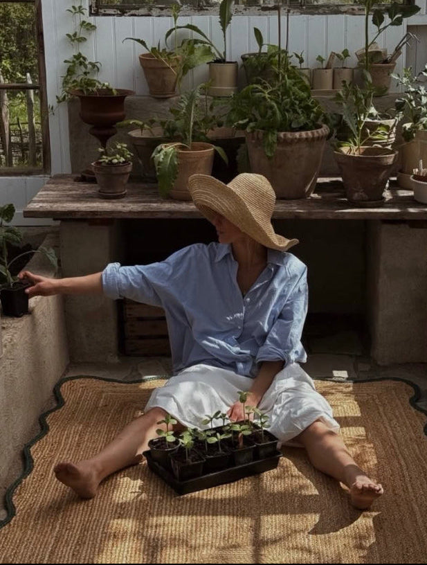 A woman wearing a wide-brimmed straw hat, a light blue button-up shirt, and white shorts sits barefoot on a woven mat inside a sunlit greenhouse, holding a tray of small potted plants. Shelves behind her are filled with potted houseplants.