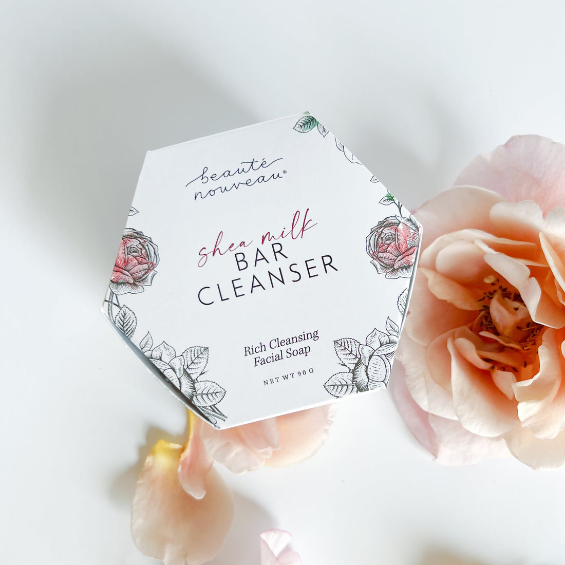 A hexagonal white box of shea milk bar cleanser, styled on a white surface with soft peach roses around it.