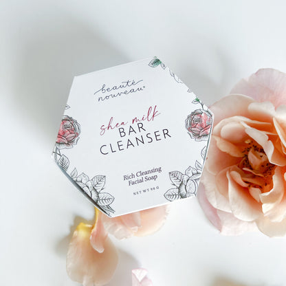 A hexagonal white box of shea milk bar cleanser, styled on a white surface with soft peach roses around it.