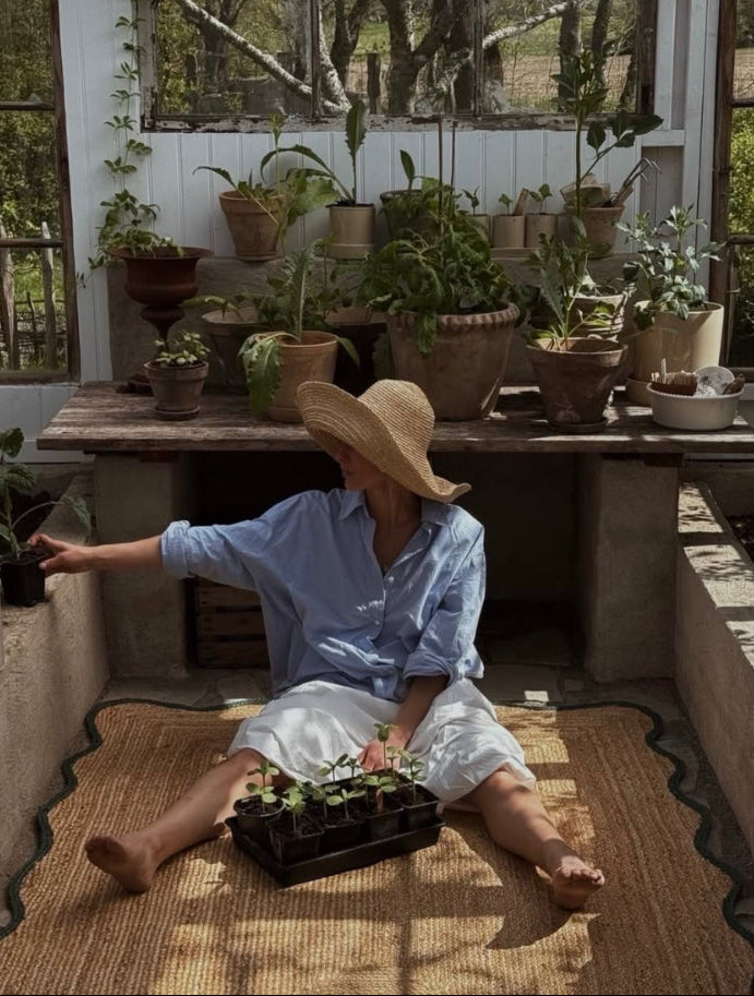 A woman wearing a wide-brimmed straw hat, a light blue button-up shirt, and white shorts sits barefoot on a woven mat inside a sunlit greenhouse, holding a tray of small potted plants. Shelves behind her are filled with potted houseplants.