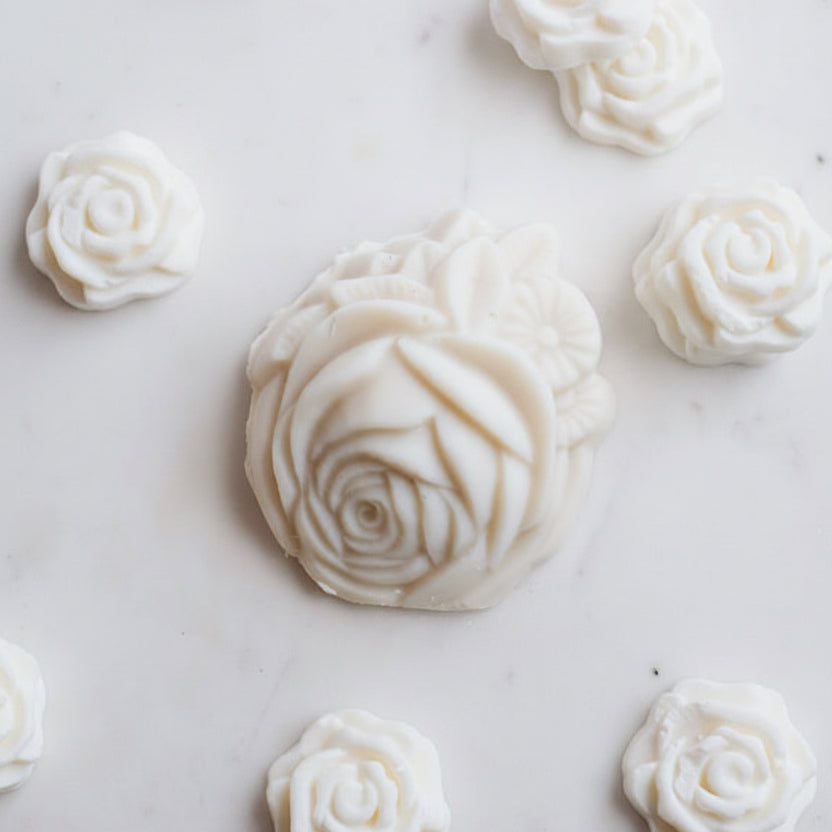 A styled flat-lay of Shea Milk Bar Facial Soap showing several sculpted off-white rose-shaped soaps on a white marble surface.